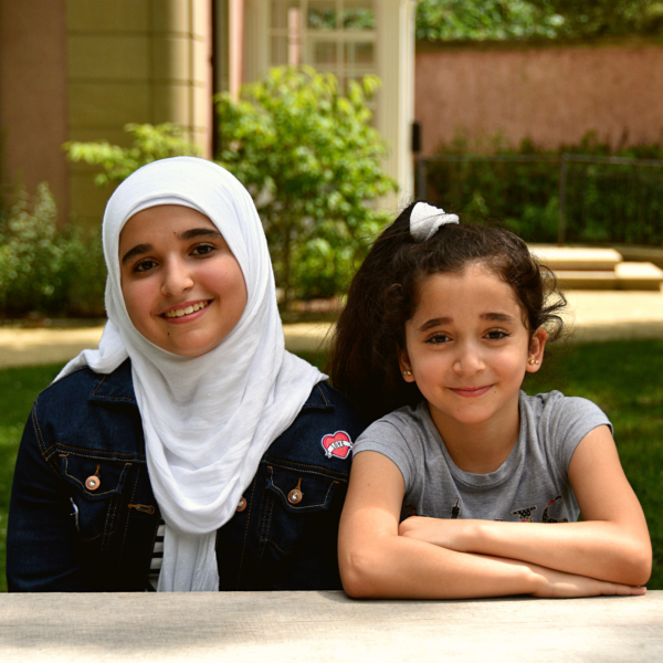 Two young Syrian refugees sit side by side in a grassy yard, smiling at the camera. One of the girls is wearing a white hijab. The other sports a high ponytail.